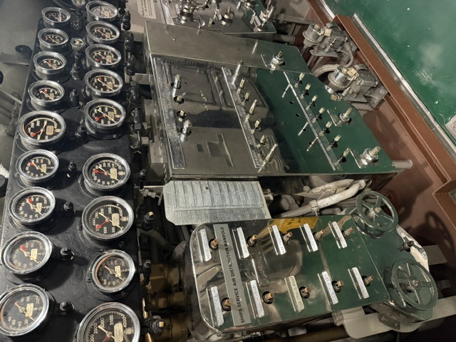 A bank of pressure gauges sits above a polished ship's service air distribution manifold with numerous valves and handwheels in a machinery space aboard USS Alabama.