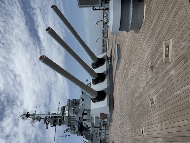 View along the wooden forward main deck of USS Alabama showing a triple 16-inch main battery gun turret with its guns elevated, and the ship's superstructure and mast beyond.