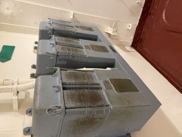 Three gray-painted electrical resistor/enclosure units mounted on a white bulkhead above a red deck, with overhead piping and a green marker plate at upper right.