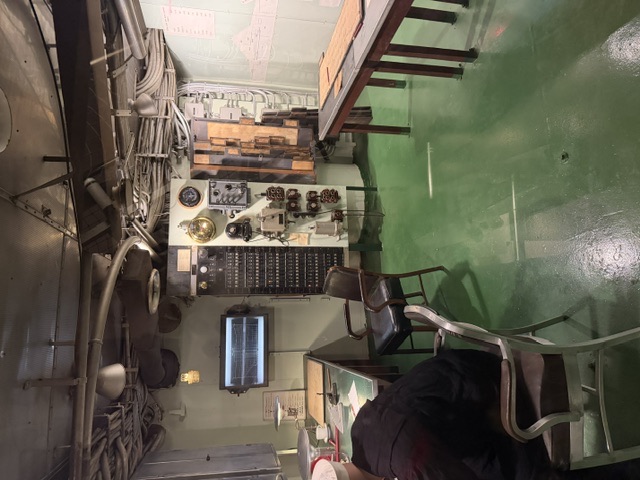 An interior plotting/control room aboard USS Alabama showing a central instrument and telephone status board, wooden plotting tables, a display monitor, and a seated crew mannequin on a green-painted deck.