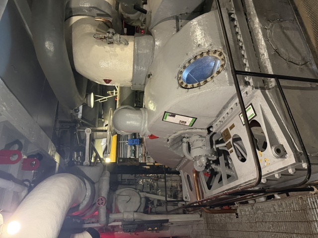 A large propulsion-machinery turbine casing in an engine room aboard USS Alabama, with white lagged (insulated) steam piping running overhead and along the bulkhead.