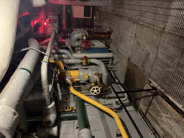 A lower-level machinery space aboard USS Alabama showing insulation-lagged overhead steam piping, color-coded pipe runs, and several handwheel valves above a metal grating deck, lit in part by a red compartment light.