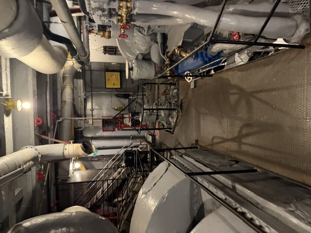 View down the central walkway of a USS Alabama engine room showing large gray-painted turbine/reduction-gear casings, brass throttle and valve stations, and insulated overhead piping.