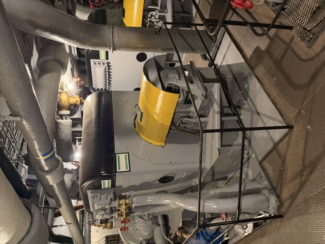 A large gray-and-yellow steam turbine machinery unit sits behind a black safety railing in an engine room aboard USS Alabama, with insulated (lagged) steam piping running overhead.