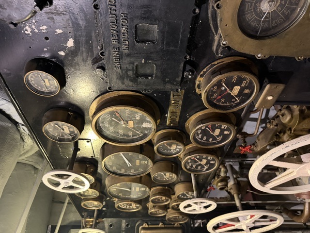 An instrument gauge board in the engine room of USS Alabama (BB-60), showing the cast 'Engine Revolutions Indicator' and 'No. 1 Unit' markings above banks of pressure/RPM dials and valve handwheels, with lagged overhead insulation at upper left.