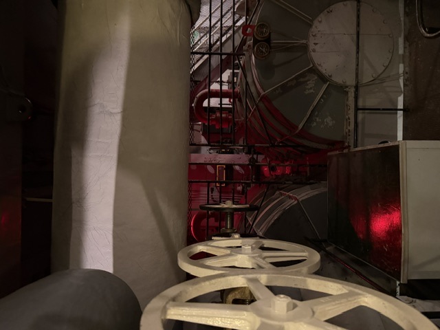 View of a USS Alabama fireroom boiler front showing valve handwheels, circular boiler drum access hatches, red steam piping, and a large asbestos-lagged overhead steam pipe.