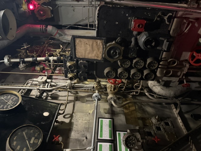 The front of a ship's boiler aboard USS Alabama, showing the circular fuel-oil burner register ports, a central furnace sight port, steam pressure gauges, and hand-operated valves, with green museum placards mounted on the adjacent bulkhead.