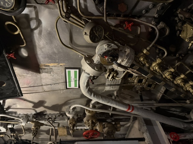 Photo shows the front of a boiler in a USS Alabama fireroom, with a white cloth-lagged steam pipe crossing an array of red-handwheel valves, pressure gauges, and brass manifold fittings.