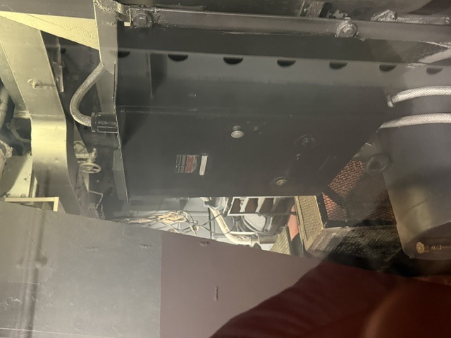 A dimly lit machinery compartment aboard USS Alabama showing a large bolted metal enclosure with an unreadable nameplate, adjacent mesh-fronted cabinet, and overhead piping and conduit.