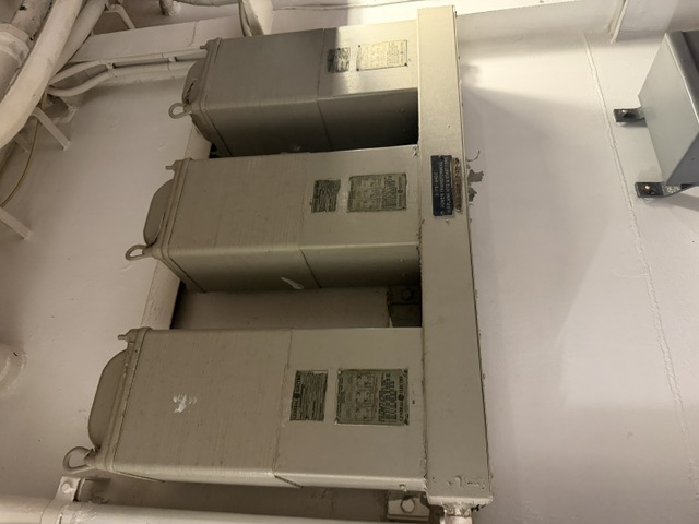 Three General Electric transformers bolted to a bulkhead aboard USS Alabama, wired as a three-phase power transformer bank with overhead cable runs.