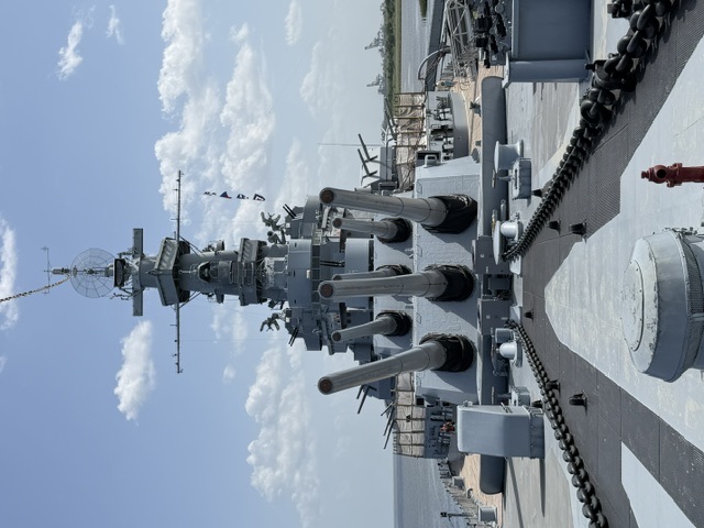 View from the forward main deck of USS Alabama looking aft at the two superimposed forward 16-inch triple gun turrets, superstructure, and mast under a partly cloudy sky.