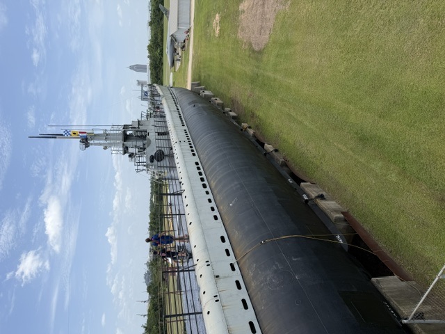Exterior view of the museum-mounted USS Drum submarine showing the port side of the hull, main deck walkway with visitors, and the conning tower against a clear sky.