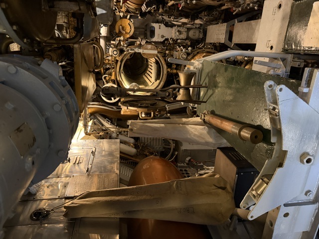 Interior view aboard USS Drum looking through an open circular watertight door between compartments, with a copper-colored tank, hanging canvas covers, and assorted valves, piping and machinery.