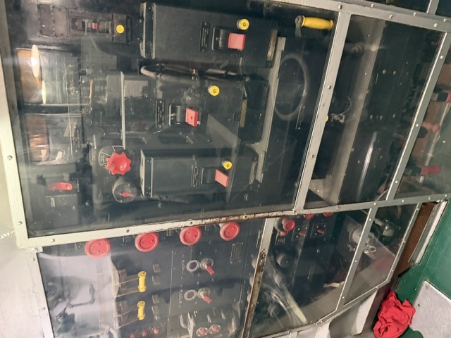 An electrical control switchboard aboard USS Drum, featuring rows of black metal panels with circuit breakers, rotary control handles, and red-capped switches, enclosed behind a clear protective barrier for museum visitors.