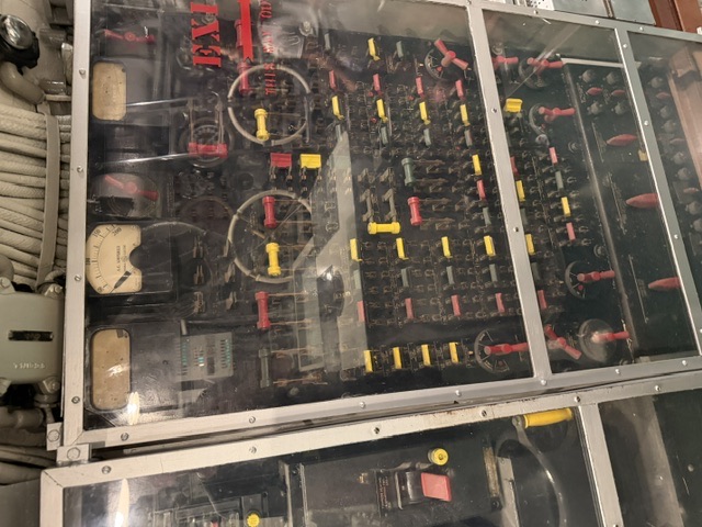 An electrical maneuvering switchboard aboard USS Drum showing meters, handwheels, and banks of red and yellow control levers behind a museum plexiglass cover, with lagged overhead piping visible at the top.