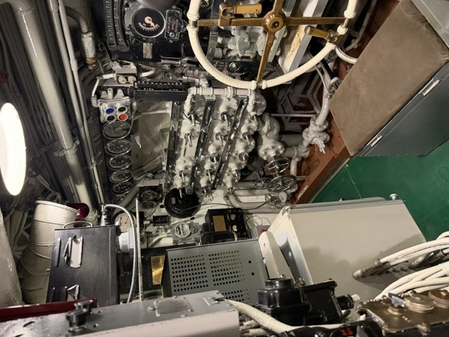 A view inside an engine room aboard USS Drum showing the top of a main diesel engine with rows of cylinder-head covers, an overhead gauge panel, and white lagged (insulated) piping running to a large brass-fitted valve.