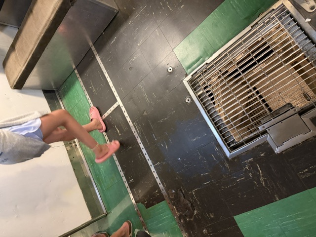 A visitor in sandals walks across the tiled interior deck of USS Drum past a recessed metal floor grating and a wall-mounted bench.