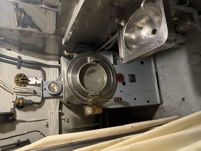 A stainless-topped galley pressure cooker with a round locking lid and pressure gauge sits beside a small stainless steel sink in a galley/scullery space aboard USS Drum.