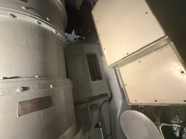 A large gray-painted cylindrical piece of machinery with bolted flanges and an illegible builder's nameplate sits above cream-colored deck panels in a below-decks space aboard USS Drum.