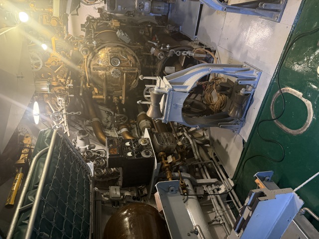 Interior view of the torpedo room aboard USS Drum showing the bronze torpedo tube breech doors with their valve manifolds and pressure gauges alongside a blue-painted torpedo handling skid.