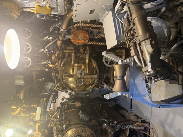 Interior view of the torpedo room aboard USS Drum (SS-228), showing the round bronze torpedo tube breech doors and associated loading equipment with overhead piping.