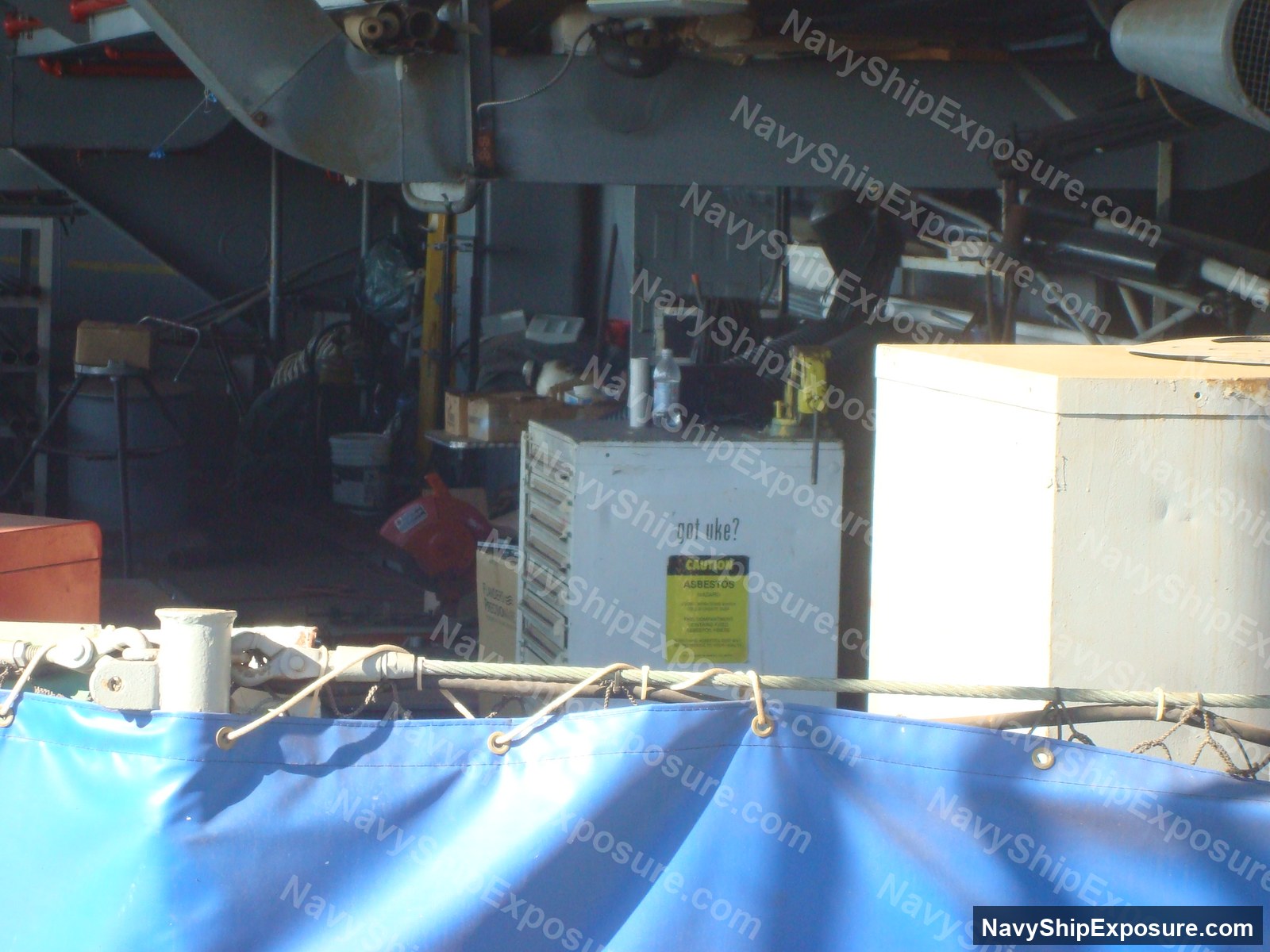 USS Midway — Interior view of a ship's machinery space showing industrial equipment, piping, and a container labeled 'got uke?' with an asbestos caution warning label visible on the bulkhead.