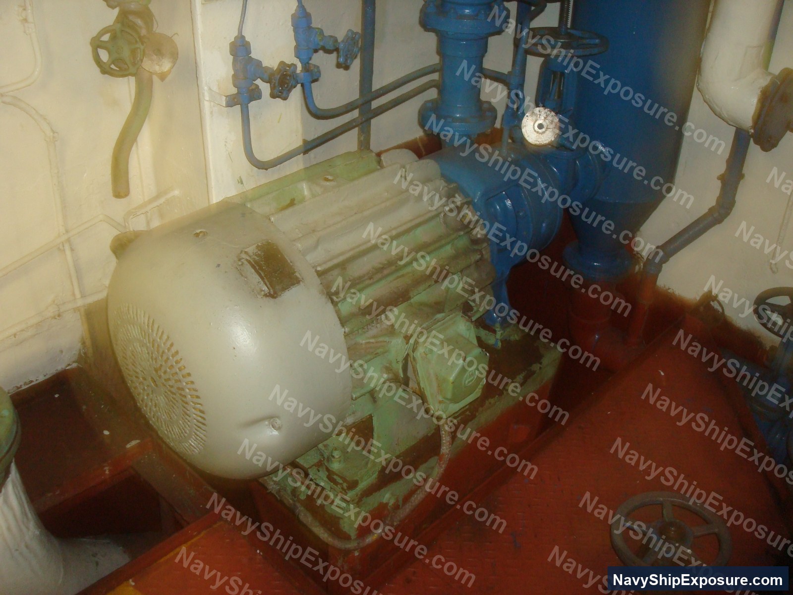 USS Midway — Engine room or machinery space aboard USS Midway showing a large electric motor on a green base mounted on red deck plating, connected to blue piping and valve systems typical of naval propulsion or auxiliary equipment.