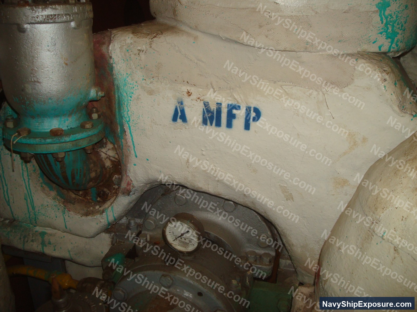 USS Midway — Engine room or machinery space aboard USS Midway showing industrial piping with turquoise corrosion deposits, a pressure gauge, and stenciled marking 'A MFP' on deteriorated cream-colored surfaces typical of 1940s-era naval vessel construction.