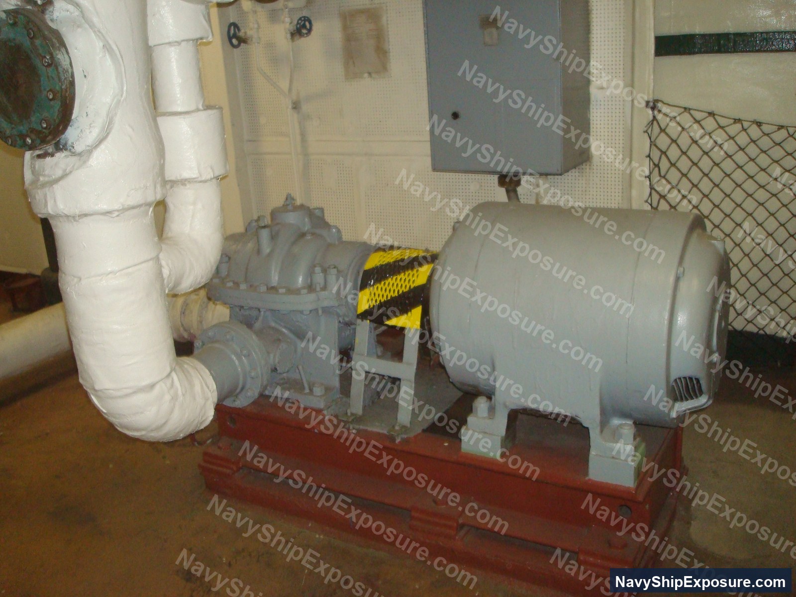 USS Midway — Engine room or machinery space equipment showing a centrifugal pump with electric motor mounted on a red base, with white insulated piping and yellow/black caution striping, typical of USS Midway's engineering spaces from the 1945-1992 service era.