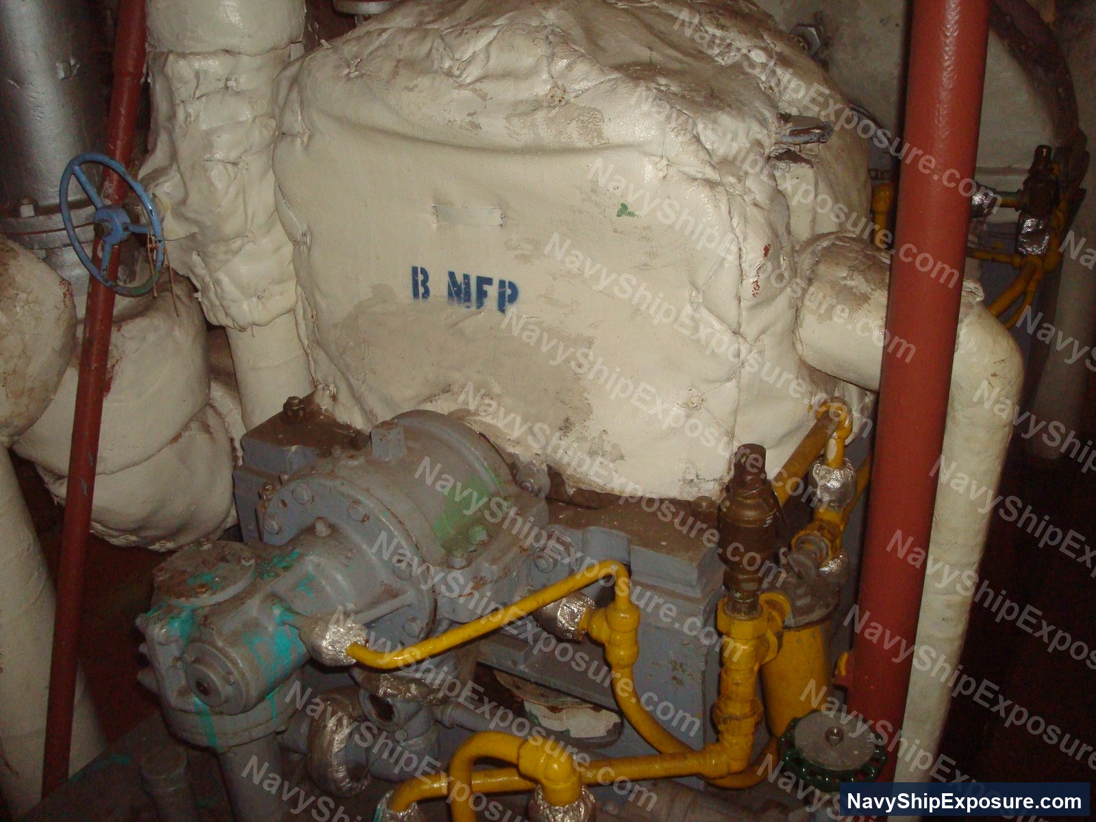 USS Midway — Engine room or machinery space aboard USS Midway showing insulated piping and equipment with visible thermal insulation material (likely containing asbestos), yellow valve handles, and red structural supports.